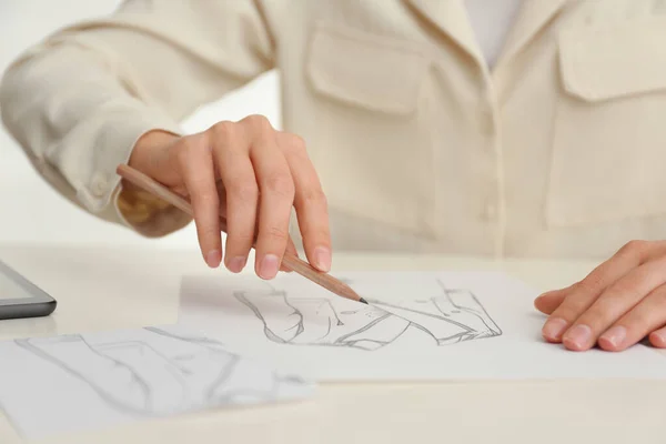 Woman drawing clothes with pencil on sheet of paper at white table, closeup