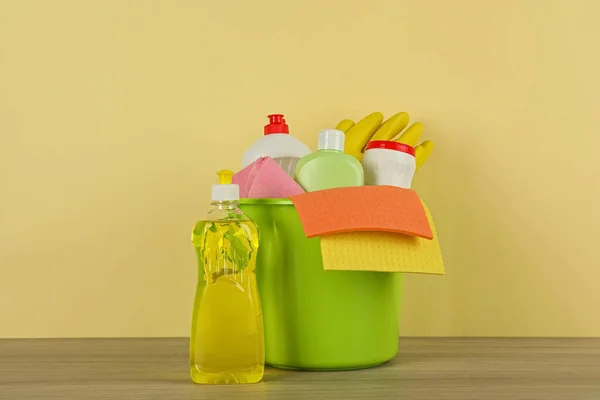 Bucket with different cleaning supplies on wooden floor near beige wall