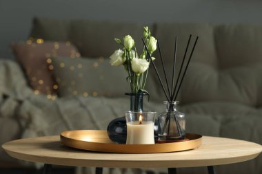 Burning candle with oil reed diffuser and bouquet on wooden table in living room