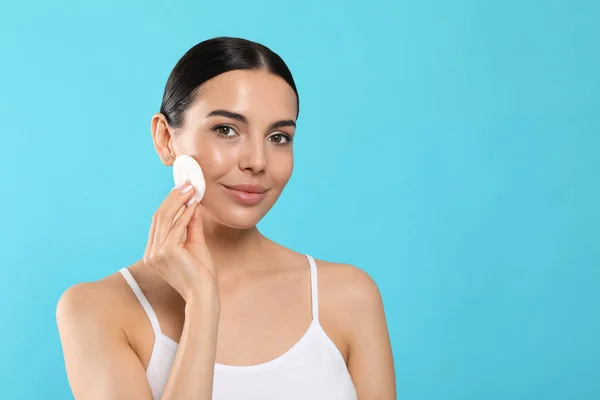 Young woman using cotton pad with micellar water on light blue background, space for text