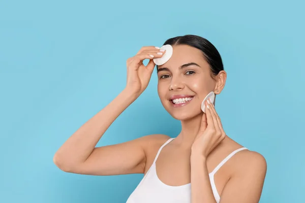 Young woman using cotton pads with micellar water on light blue background