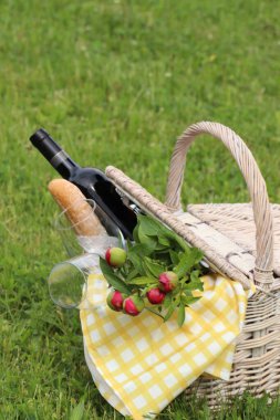 Picnic basket with wine, bread and flowers on green grass outdoors