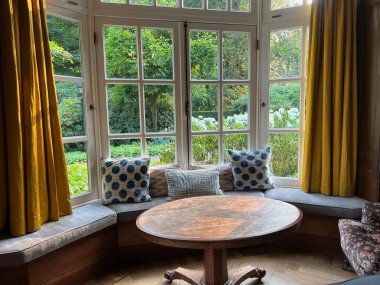 Wooden table near cozy bay window seat with cushions in room. Interior design