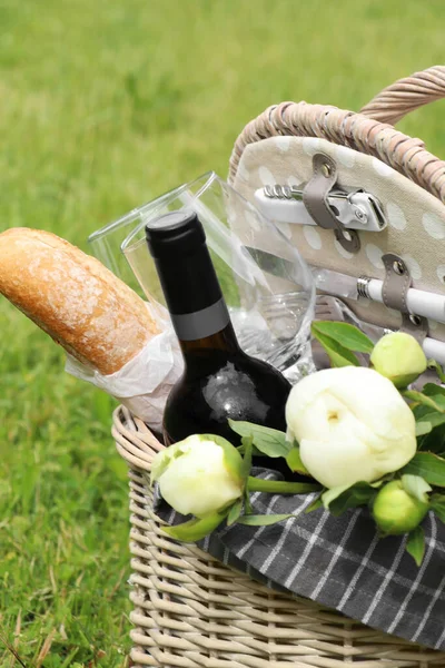 Picnic basket with wine, bread and flowers on green grass outdoors