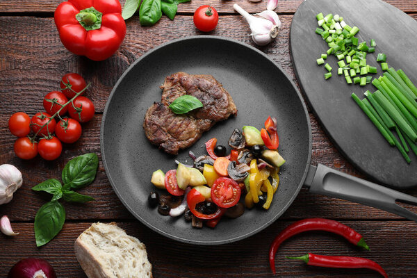 Tasty fried steak with vegetables in pan and ingredients on wooden table, flat lay