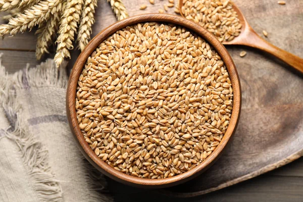 Wheat grains in bowl, spoon and spikes on wooden table, flat lay