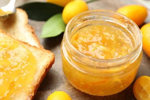 Delicious kumquat jam in jar, tasty toast and fresh fruits on wooden board, closeup