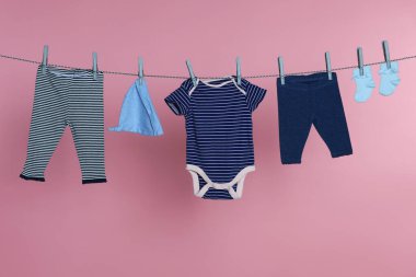 Different baby clothes drying on laundry line against pink background