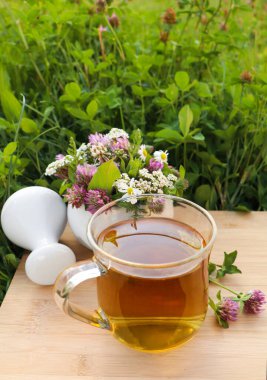 Cup of aromatic herbal tea, pestle and ceramic mortar with different wildflowers on wooden board in meadow