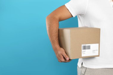 Courier holding cardboard box on light blue background, closeup. Space for text