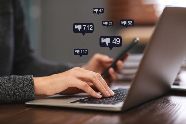 Closeup view of young woman using modern laptop with smartphone indoors and virtual dislike icons at table. Cyberbullying concept