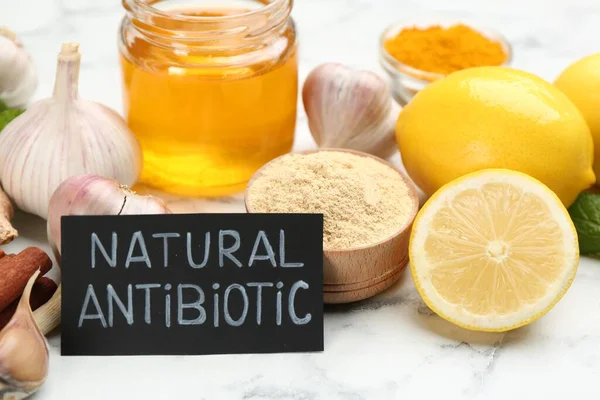 Different fresh products and card with phrase Natural Antibiotic on white marble table, closeup