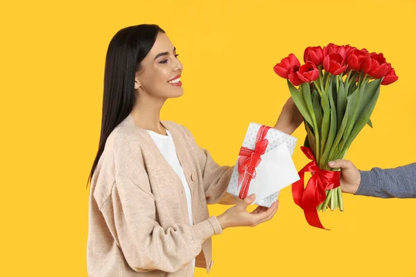 黄色の背景に男から赤いチューリップの花束 グリーティングカードとギフトボックスを受け取った幸せな女性 3月8日 — ストック写真
