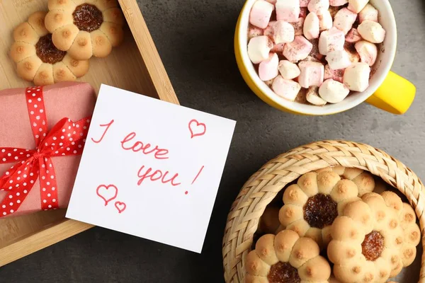 Note with phrase I love You near gift box, cookies and cup of hot drink on dark table, flat lay