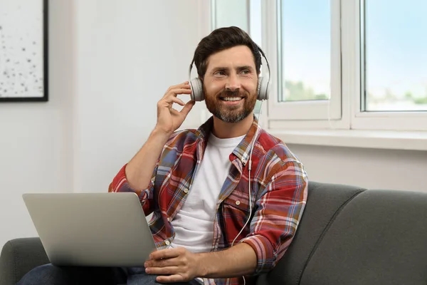 Smiling bearded man with headphones and laptop on sofa indoors