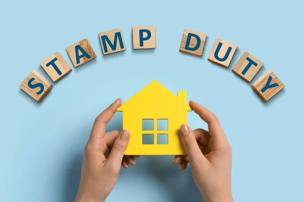 Woman holding house figure and wooden cubes with text Stamp Duty on light blue background, top view