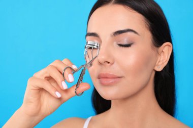 Beautiful young woman using eyelash curler on light blue background