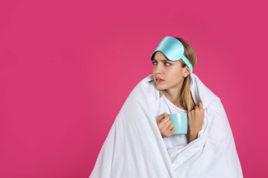 Pembe arka planda battaniyeye sarılı, uyku maskesi takmış üzgün genç bir kadın. Metin için boşluk