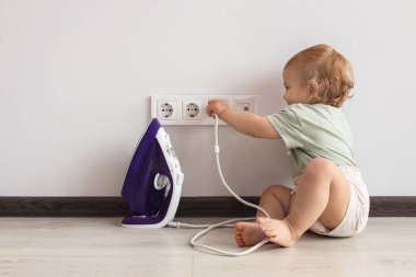 Cute baby playing with electrical socket and iron plug at home. Dangerous situation