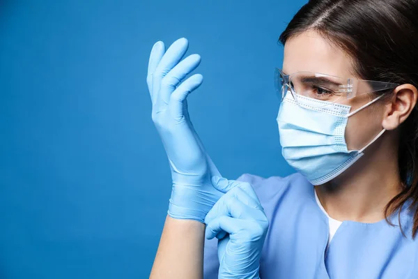 Doctor in protective mask and glasses putting on medical gloves against blue background. Space for text