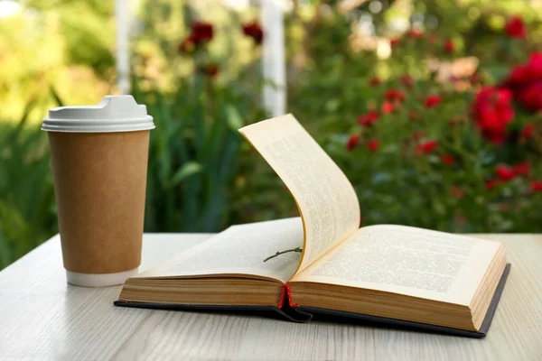Open book with paper cup of coffee on wooden table in garden