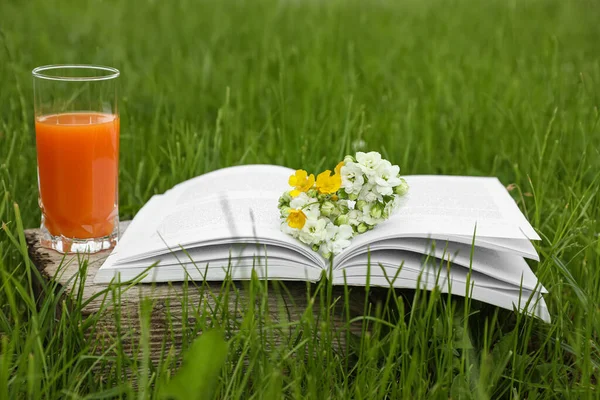 Open book with flowers and glass of juice on green grass outdoors