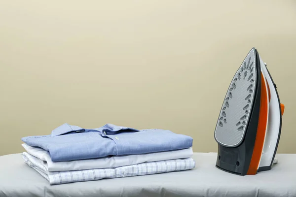 Modern iron and stack of clean clothes on board against beige background. Space for text