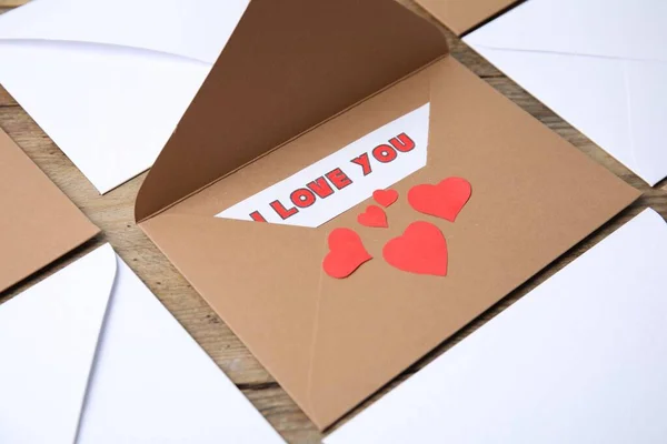 Sheet of paper with phrase I Love You, envelopes and red hearts on wooden table, closeup