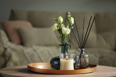 Burning candle with oil reed diffuser and bouquet on wooden table in living room