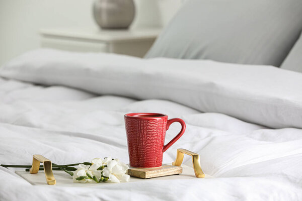 Mug of hot drink with stylish cup coaster and beautiful freesia flowers on bed in room