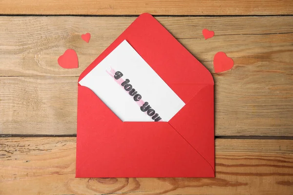 Card with phrase I Love You in envelope and paper hearts on wooden table, flat lay