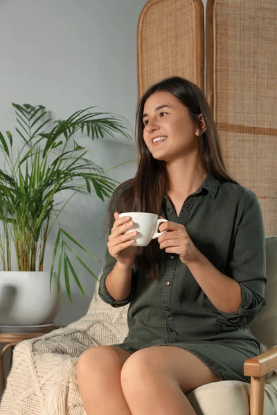 Happy young woman with cup of coffee sitting in armchair at home