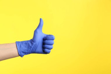 Person in medical gloves showing thumb up on yellow background, closeup of hand. Space for text