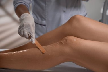 Professional cosmetologist applying gel on client's leg before laser epilation procedure in salon, closeup
