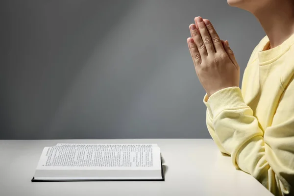 Woman praying over Bible at white table against grey background, closeup. Space for text