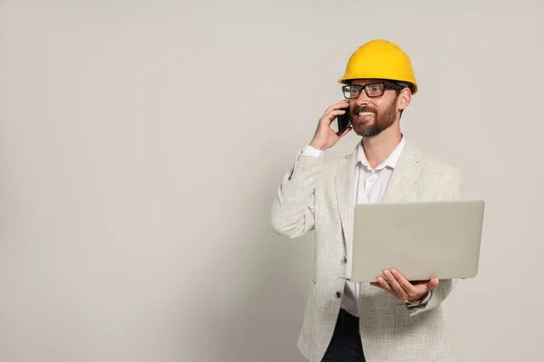 Professional engineer in hard hat with laptop talking on phone against white background, space for text