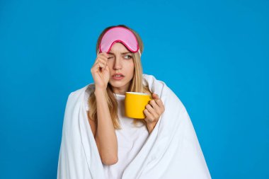 Mavi arka planda battaniyeye sarılı, uyku maskeli genç bir kadın.