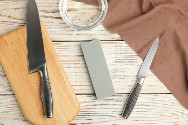 Sharpening stone, knives and water on white wooden table, flat lay