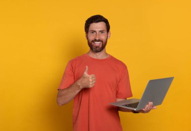 Laptoplu yakışıklı adam turuncu arka planda baş parmak işareti yapıyor.