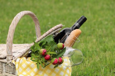 Picnic basket with wine, bread and flowers on green grass outdoors, closeup