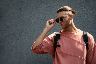 Gri duvarın yanında şık güneş gözlüklü ve sırt çantalı yakışıklı genç adam, mesaj için yer var.