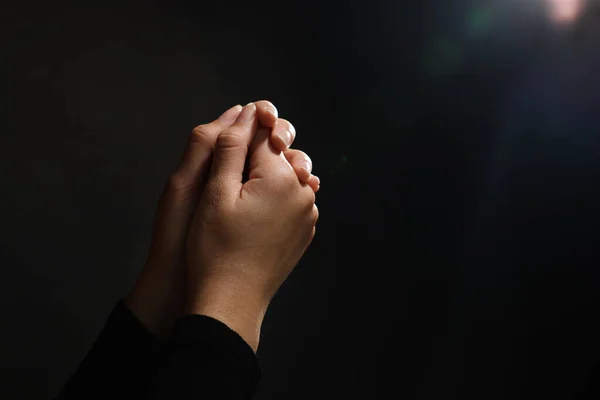 Woman holding hands clasped while praying against light in darkness, closeup. Space for text