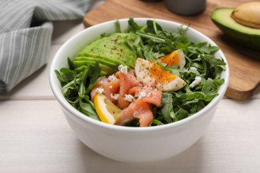 Delicious salad with boiled egg, salmon and avocado in bowl on white wooden table, closeup