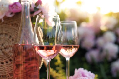 Bottle and glasses of rose wine near straw bag with beautiful peonies in garden, closeup