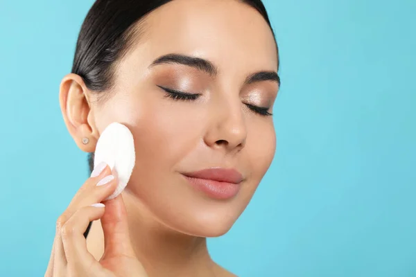 Young woman using cotton pad with micellar water on light blue background, closeup