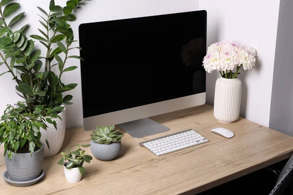 Comfortable workplace with modern computer, green houseplants and beautiful flowers on wooden table indoors