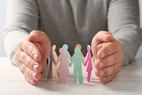 Man protecting paper human figures on white wooden table, closeup. Diversity and inclusion concept