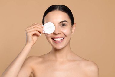 Young woman using cotton pad with micellar water on beige background
