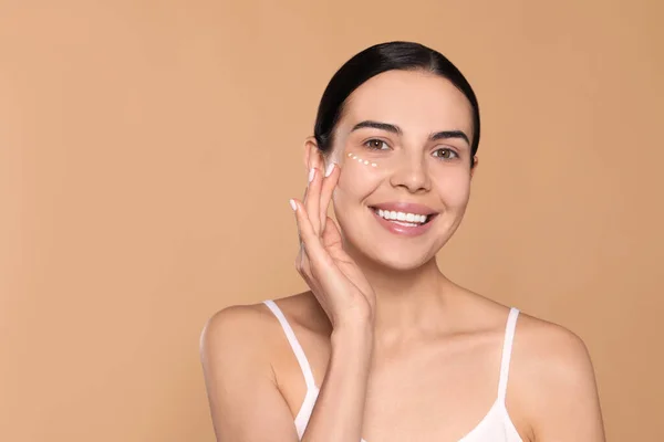 Beautiful young woman with cream on skin under eye against beige background. Space for text