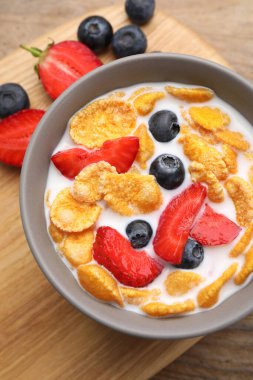 Bowl of tasty crispy corn flakes with milk and berries on wooden table, flat lay